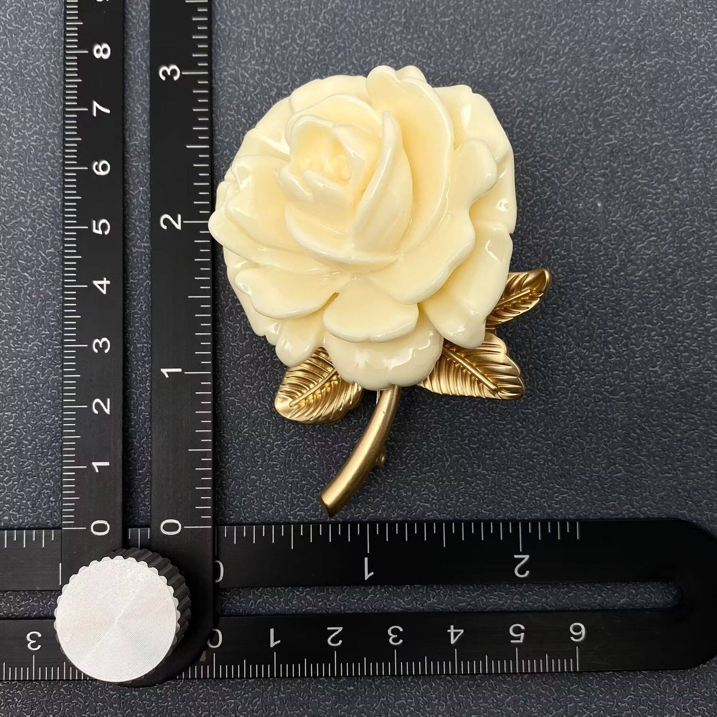 White rose brooch with gold leaves on a black surface with a ruler for scale.