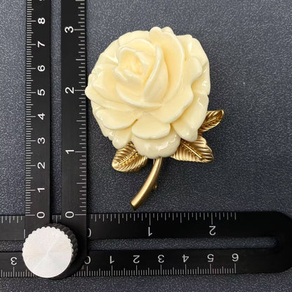 White rose brooch with gold leaves on a black surface with a ruler for scale.
