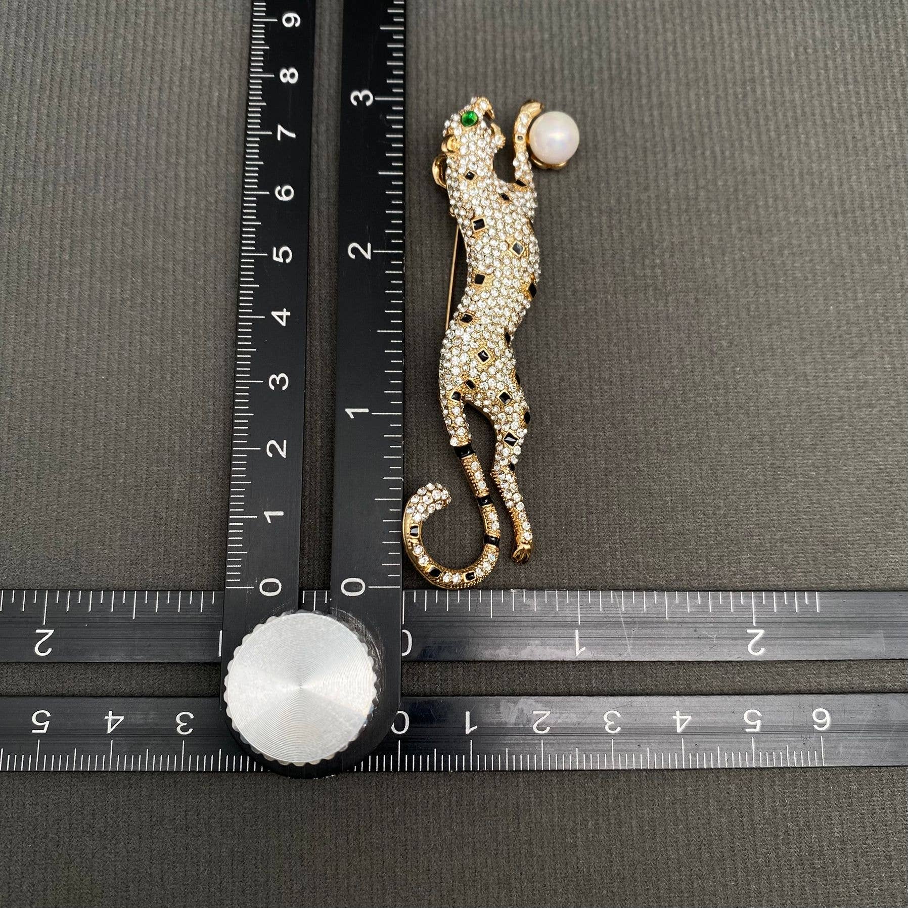 Decorative leopard brooch with gemstones next to a ruler on a gray background