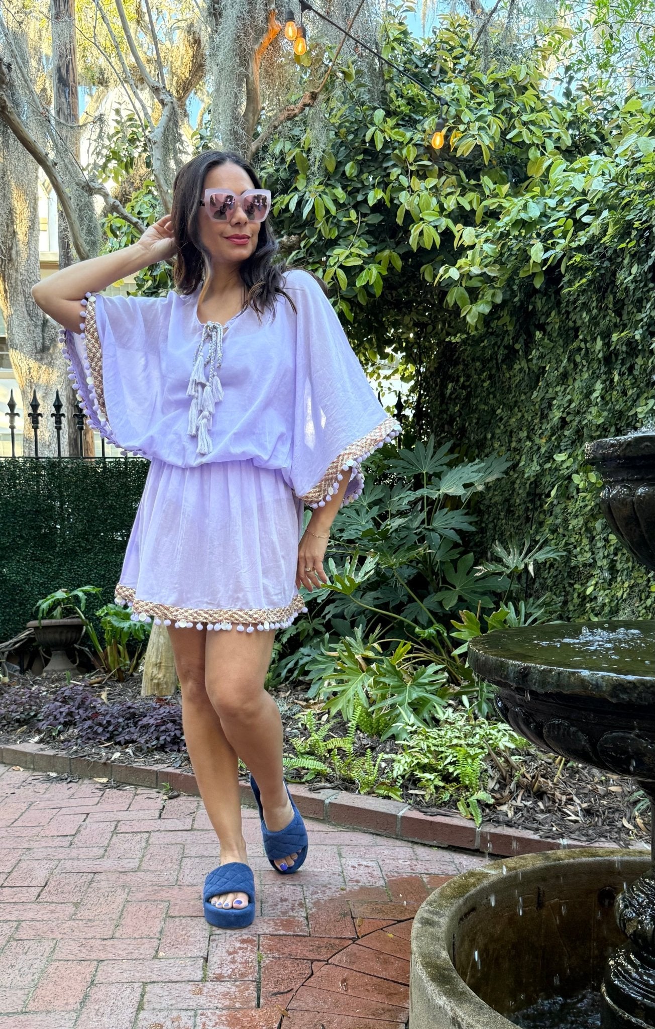 Woman in a light purple swimsuit coverup standing near a fountain with greenery in the background