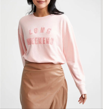 A woman wearing a light pink crewneck sweatshirt with 'LONG WEEKEND' printed in pink across the chest, paired with a brown wrap-style skirt, against a light gray studio background.