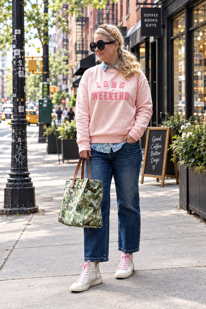 A woman stands on a city sidewalk wearing a light pink sweatshirt with 'LOVE WEEKEND' printed in pink across the chest, blue jeans, and white sneakers. She is holding a green camouflage tote bag.
