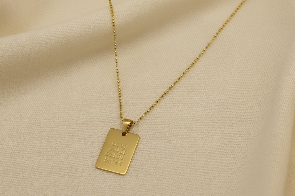 Gold necklace with a pendant that reads 'LOVE EACH OTHER MORE' on an ivory background