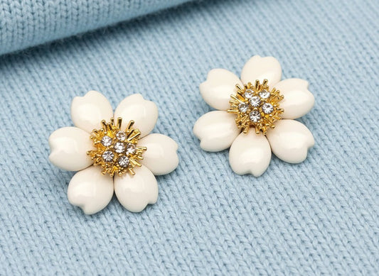 Pair of white flower-shaped earrings with gold and crystal centers on a light blue sweater material background