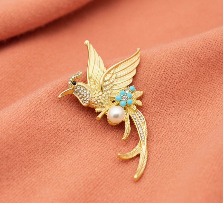 Gold hummingbird brooch with pearl and turquoise stones on a coral colored sweater fabric background