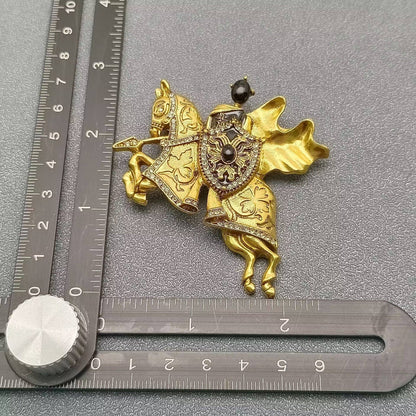 gold and black medieval knight and horse themed brooch next to a ruler for scale against a grey textured background