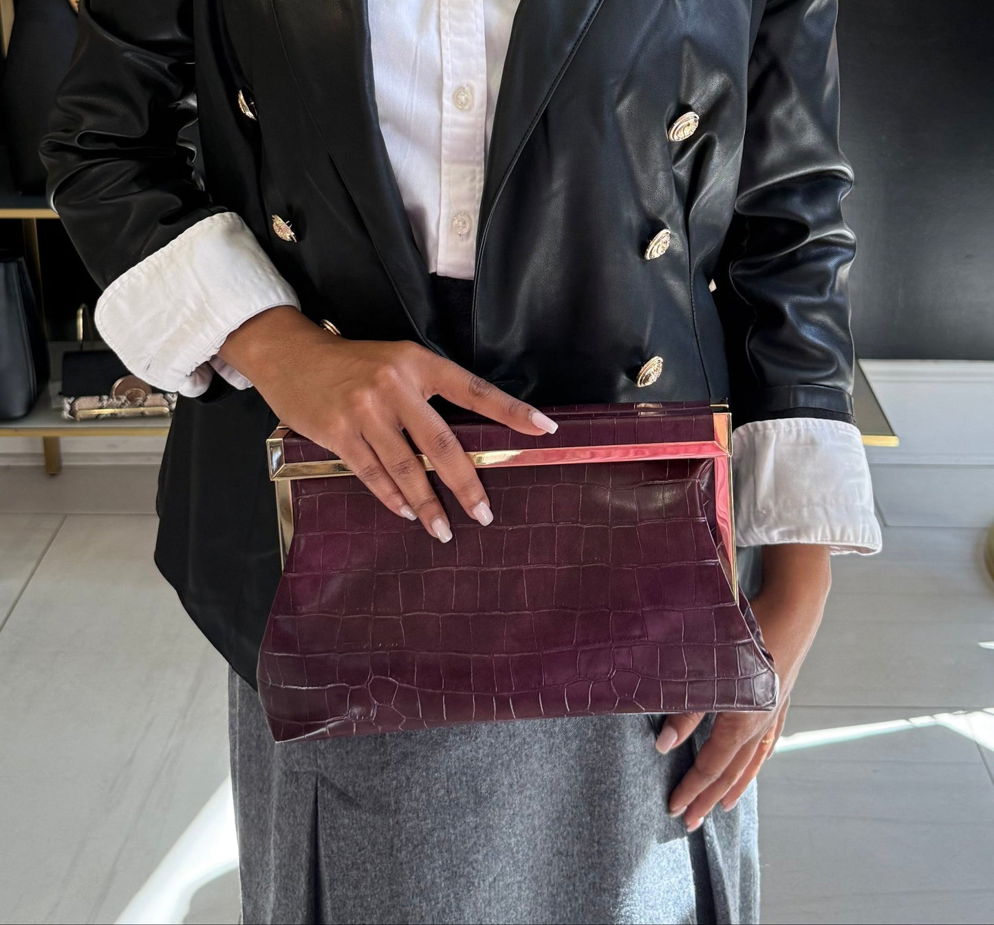 Woman holding a merlot colored croc embossed clutch in a store setting with bags and decor in the background