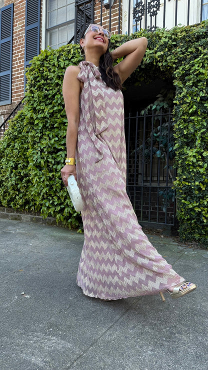 Woman in a long, chevron patterned dress standing outdoors with greenery and a building in the background.