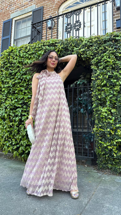 A woman wearing a metallic chevron print halter neck maxi dress with sleeveless design, standing in front of a wall with climbing plants.