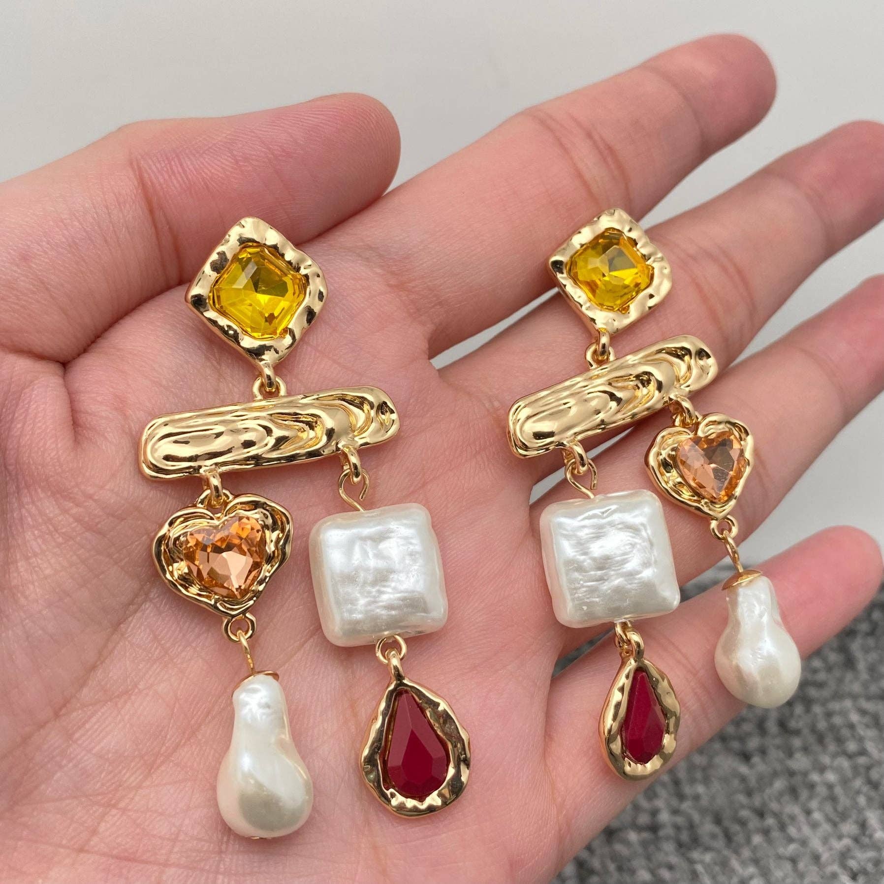 A pair of baroque style dangle earrings with geometric design, featuring gold plating, pearls, and colorful glass crystals, held in a person's hand.