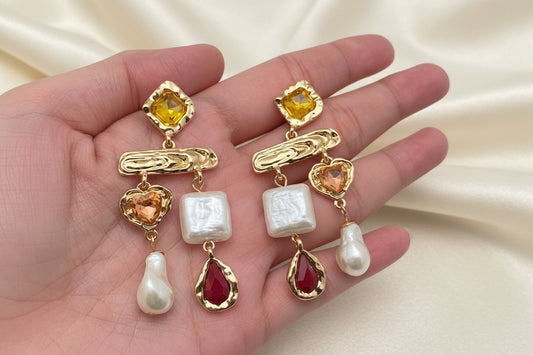 A pair of baroque style dangle earrings with geometric design, featuring gold plating, pearls, and colorful glass crystals, held in a person's hand.