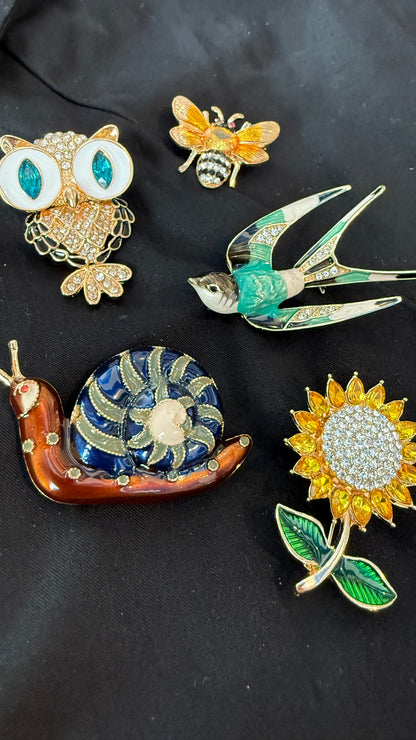 Collection of decorative pins including an owl, bee, swan, snail, and sunflower on a dark background.