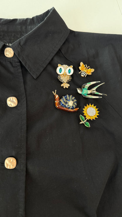Black shirt with decorative pins including an owl, bee, snail, and sunflower on a white background