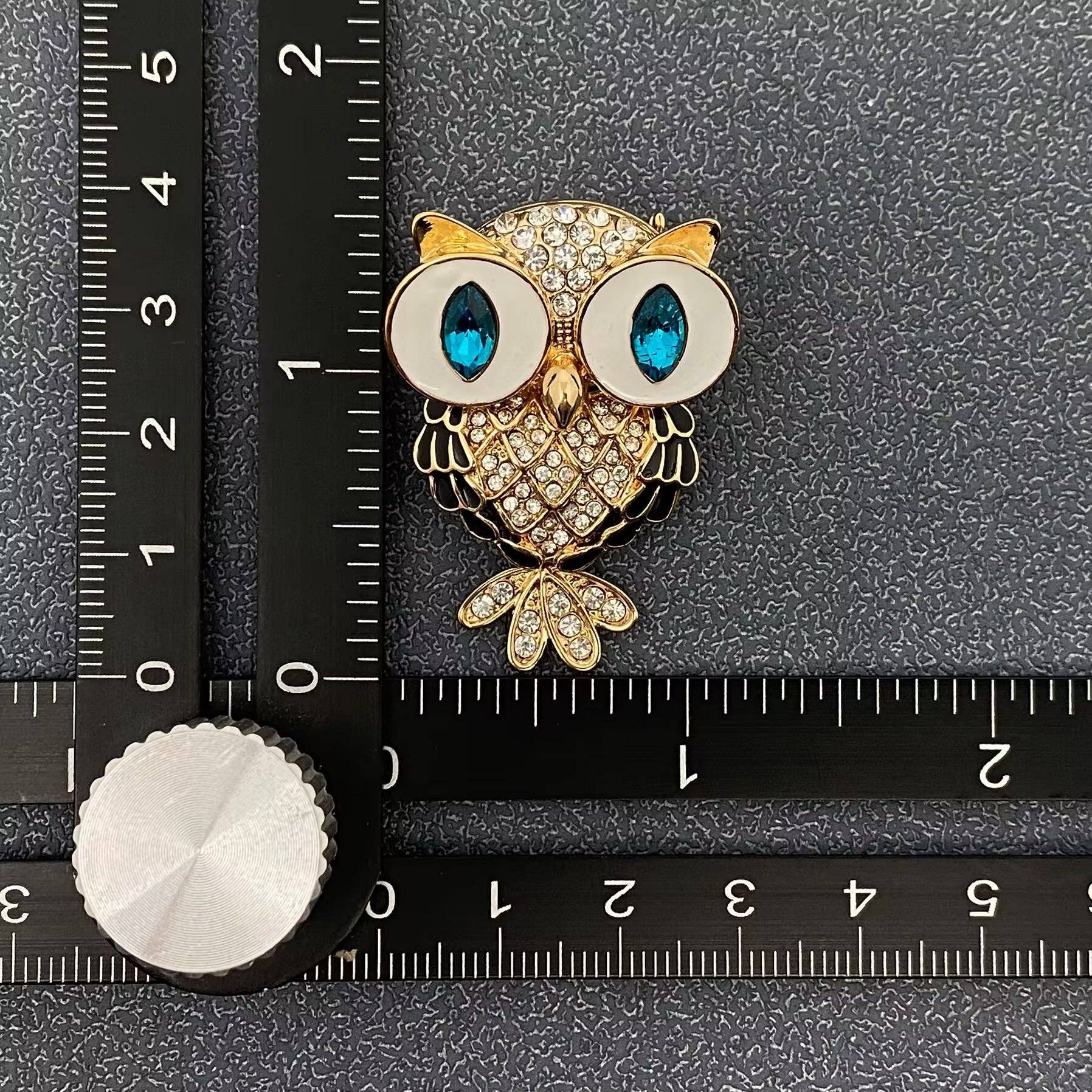 Gold owl brooch with blue eyes on a black surface with a ruler for scale.