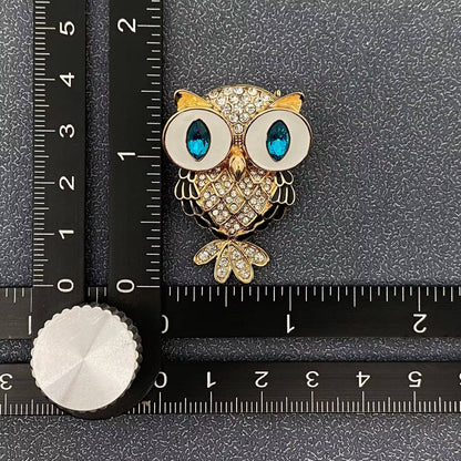 Gold owl brooch with blue eyes on a black surface with a ruler for scale.