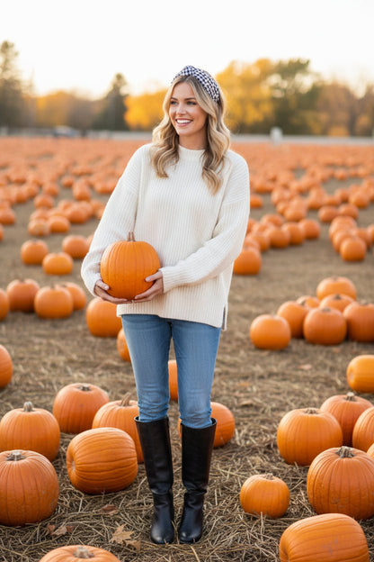 a woman wearing an ivory sweater, jeans, and a black and white gingham headband holding a pumpkin in a pumpkin patch