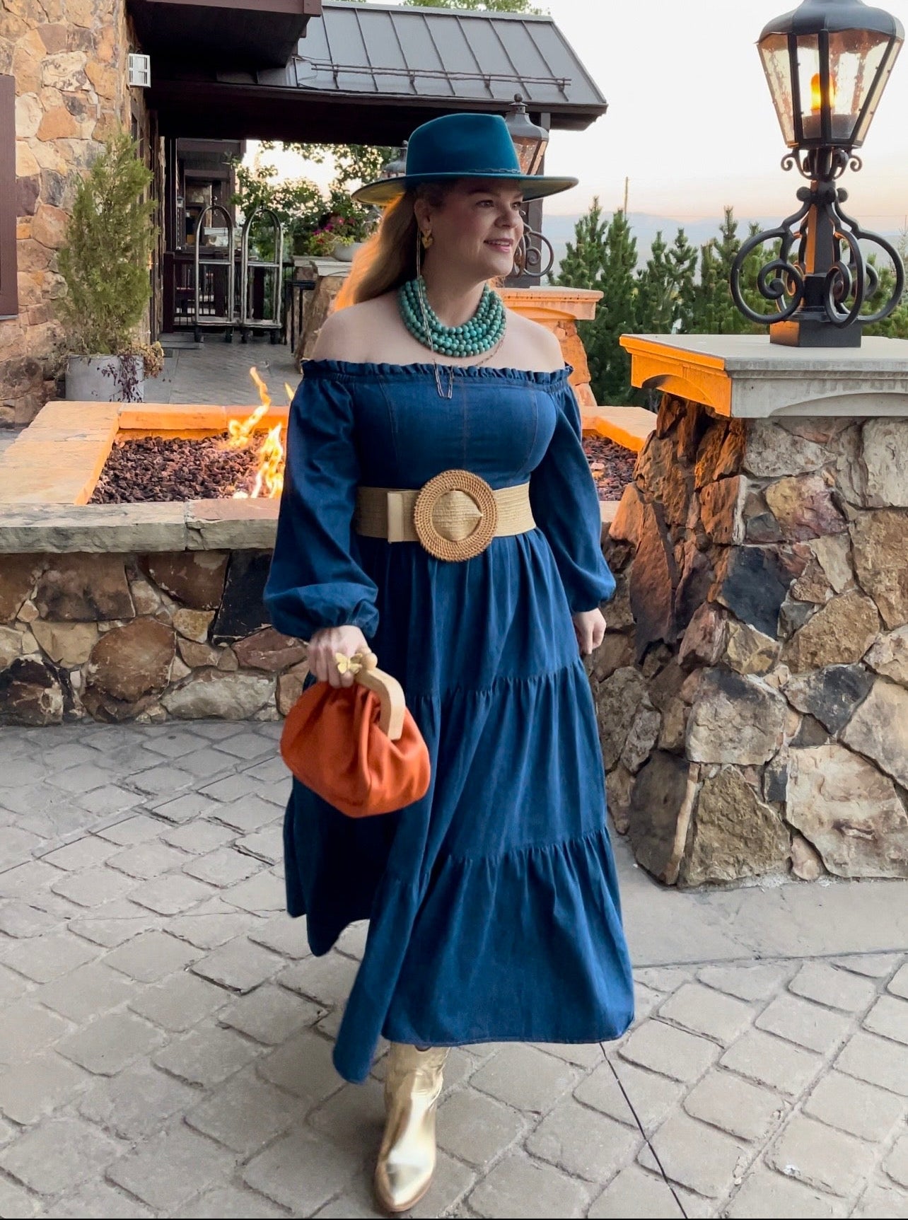 Woman in a blue denim dress with a rattan belt and orange clutch standing outdoors near a stone building.