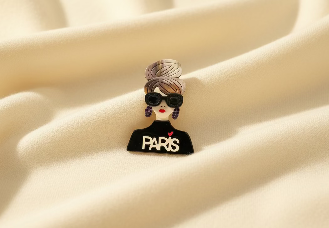 Brooch with a design of a woman wearing sunglasses and 'PARIS' text on a beige fabric background