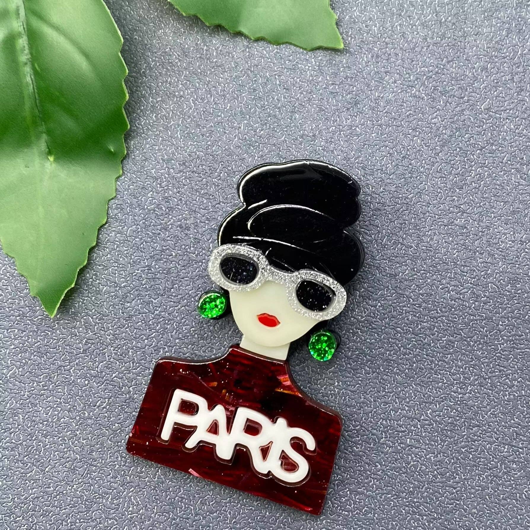 Acrylic Brooch with a stylized face wearing sunglasses and a 'PARIS' sign on a gray background with green leaves.