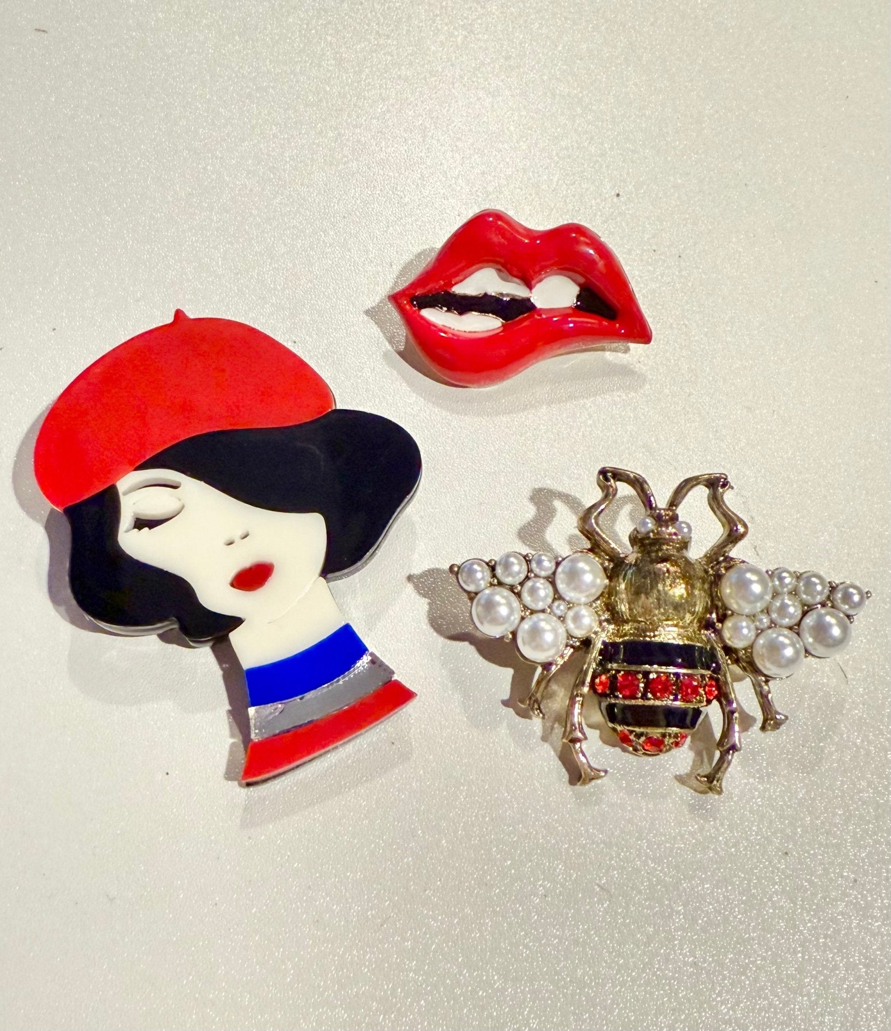 Three decorative pins: a woman with a red beret, a pair of red lips, and a bee with pearls on a light background.