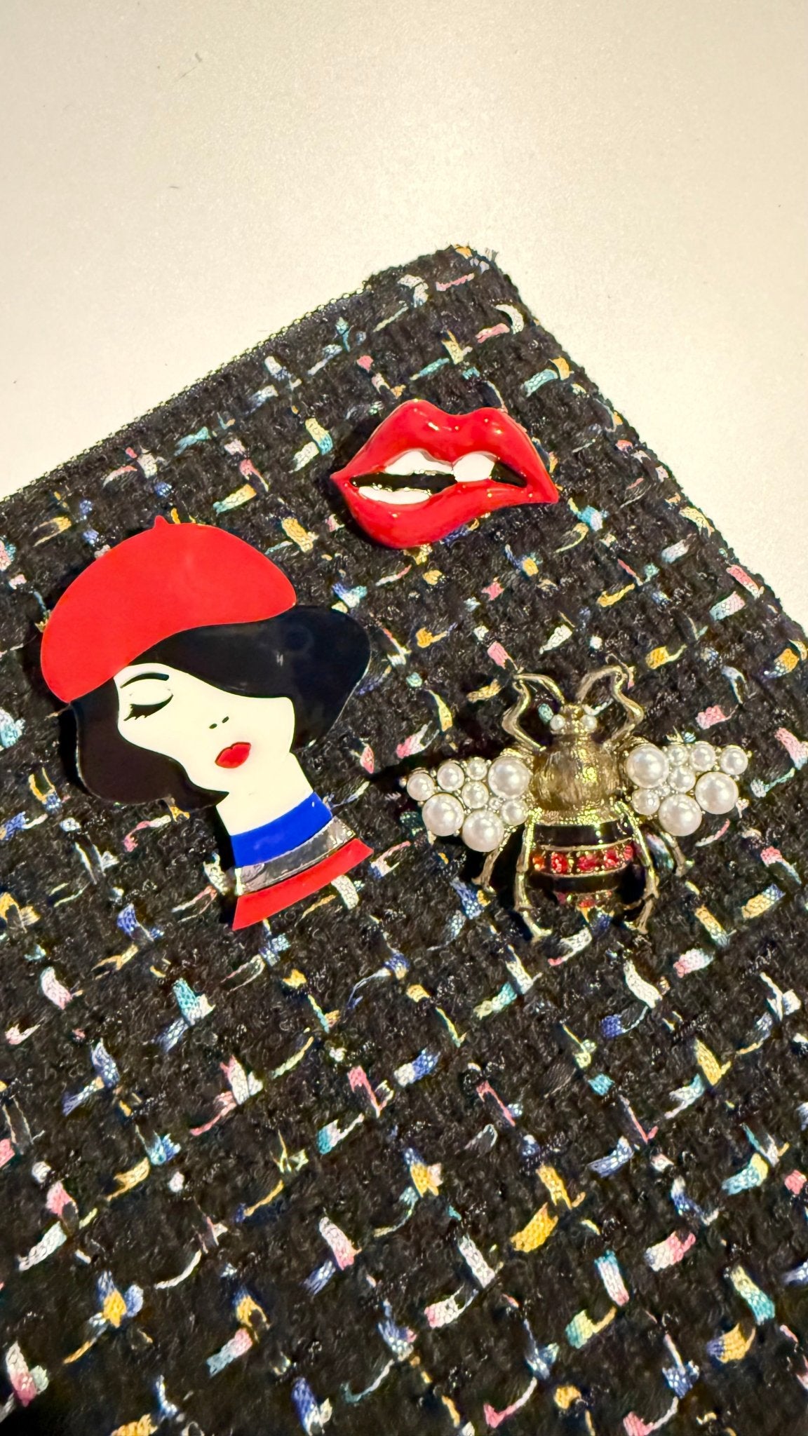 Black textured surface with decorative brooches including a woman in a red hat, red lips, and a bee.