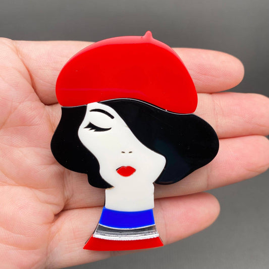 Decorative brooch with a stylized face wearing a red beret held in a hand against a gray background.