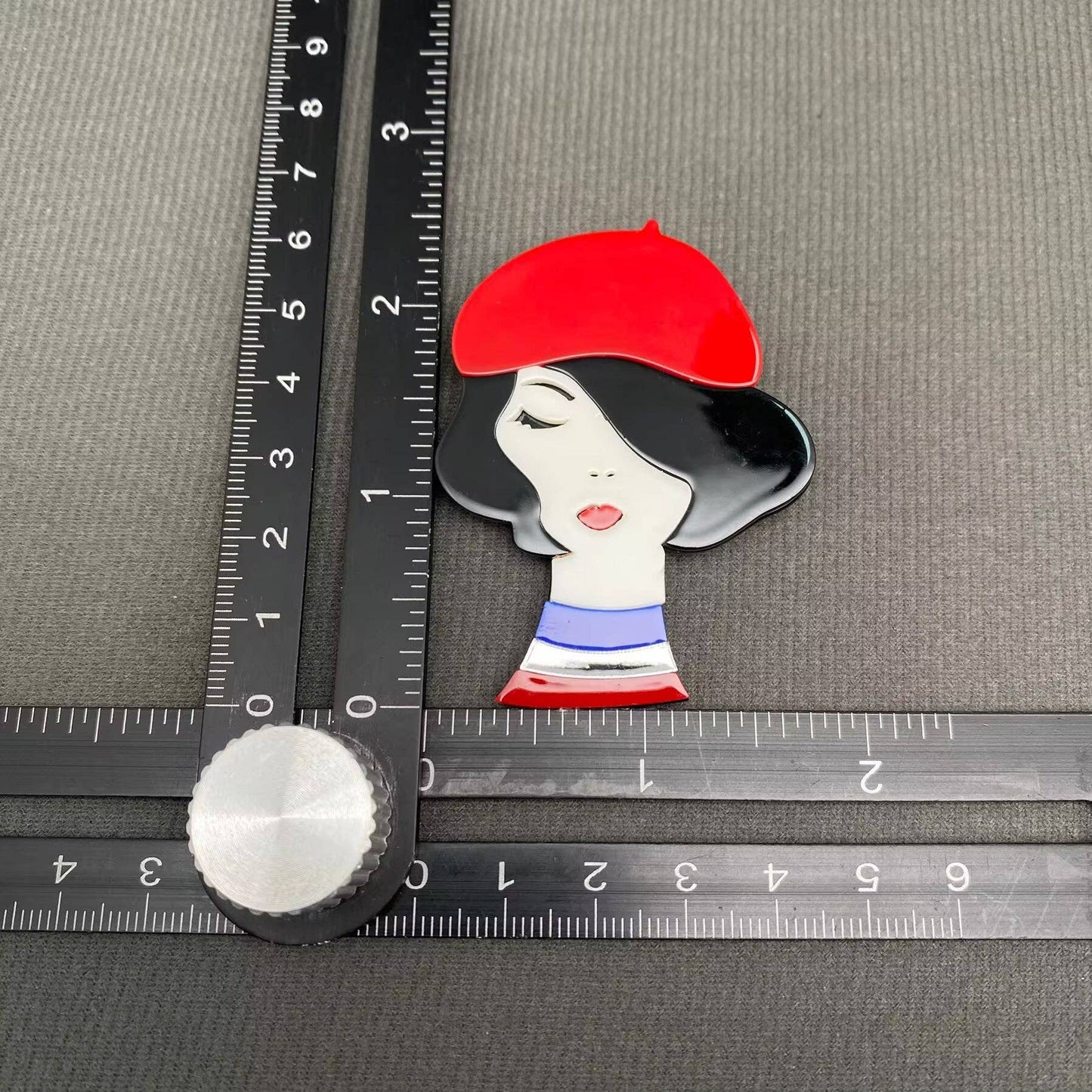 Brooch with a design of a woman's face wearing a red beret on a gray background with rulers.