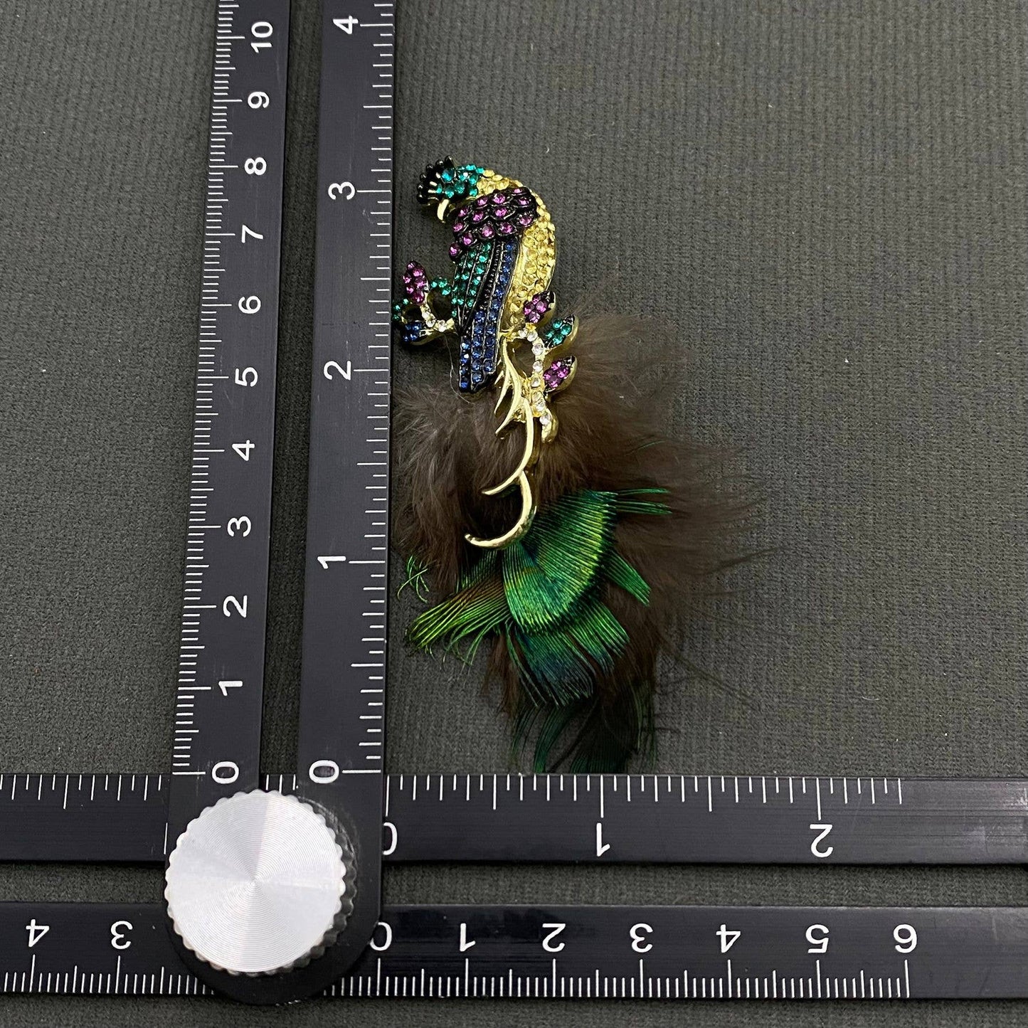 Decorative peacock brooch with feathers and gemstones next to a ruler on a gray background