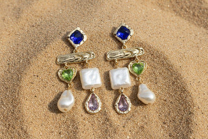 Pair of gold earrings with gemstones and pearls on a sandy background