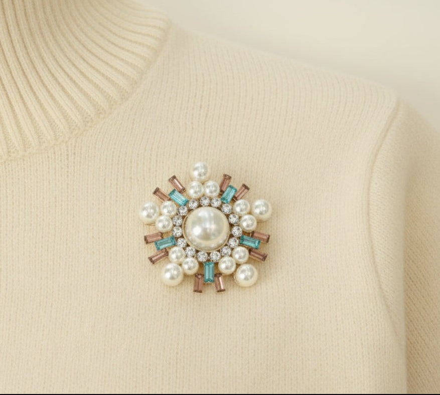 An ivory colored sweater with a decorative brooch featuring pearls and gemstones on a white background