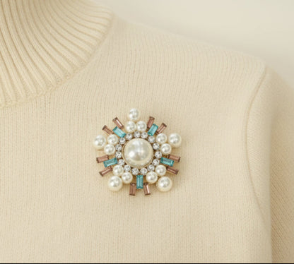 An ivory colored sweater with a decorative brooch featuring pearls and gemstones on a white background