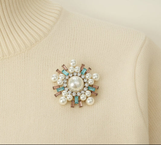 An ivory colored sweater with a decorative brooch featuring pearls and gemstones on a white background