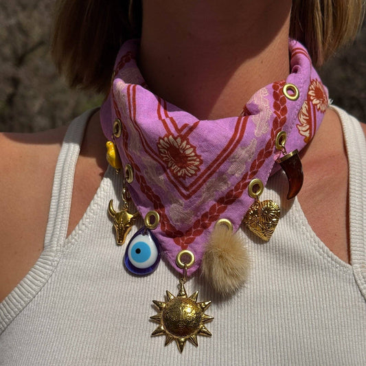 A close-up of a person wearing a pink patterned bandana-style neck scarf with gold-tone eyelets and multiple dangling charms, including a blue evil eye pendant, a gold bull charm, a gold sun pendant, and a fluffy cream pom-pom, over a white ribbed tank top.