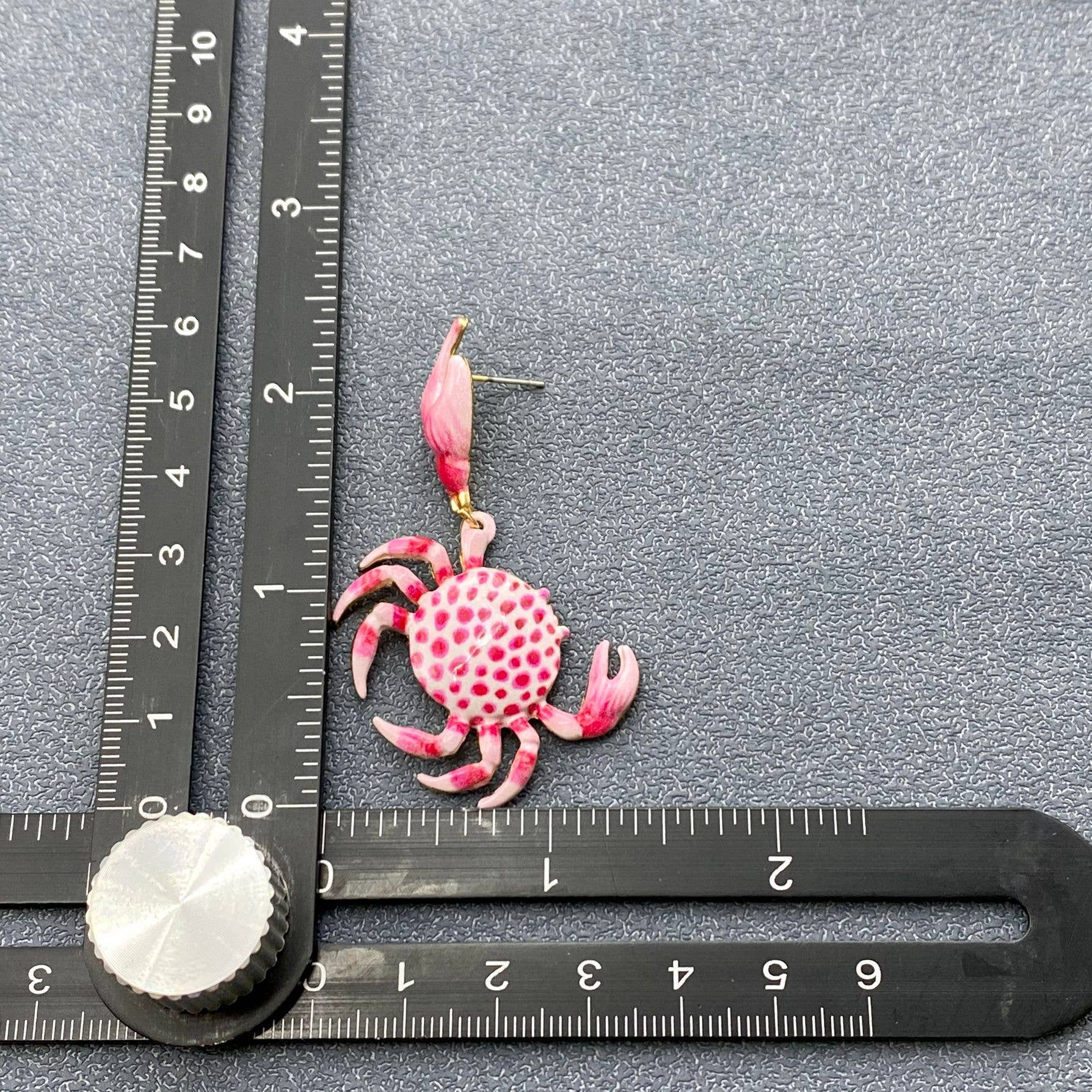 a pink and white enamel crab dangle earring next to a rule to show the size