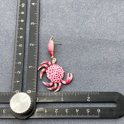 a pink and white enamel crab dangle earring next to a rule to show the size