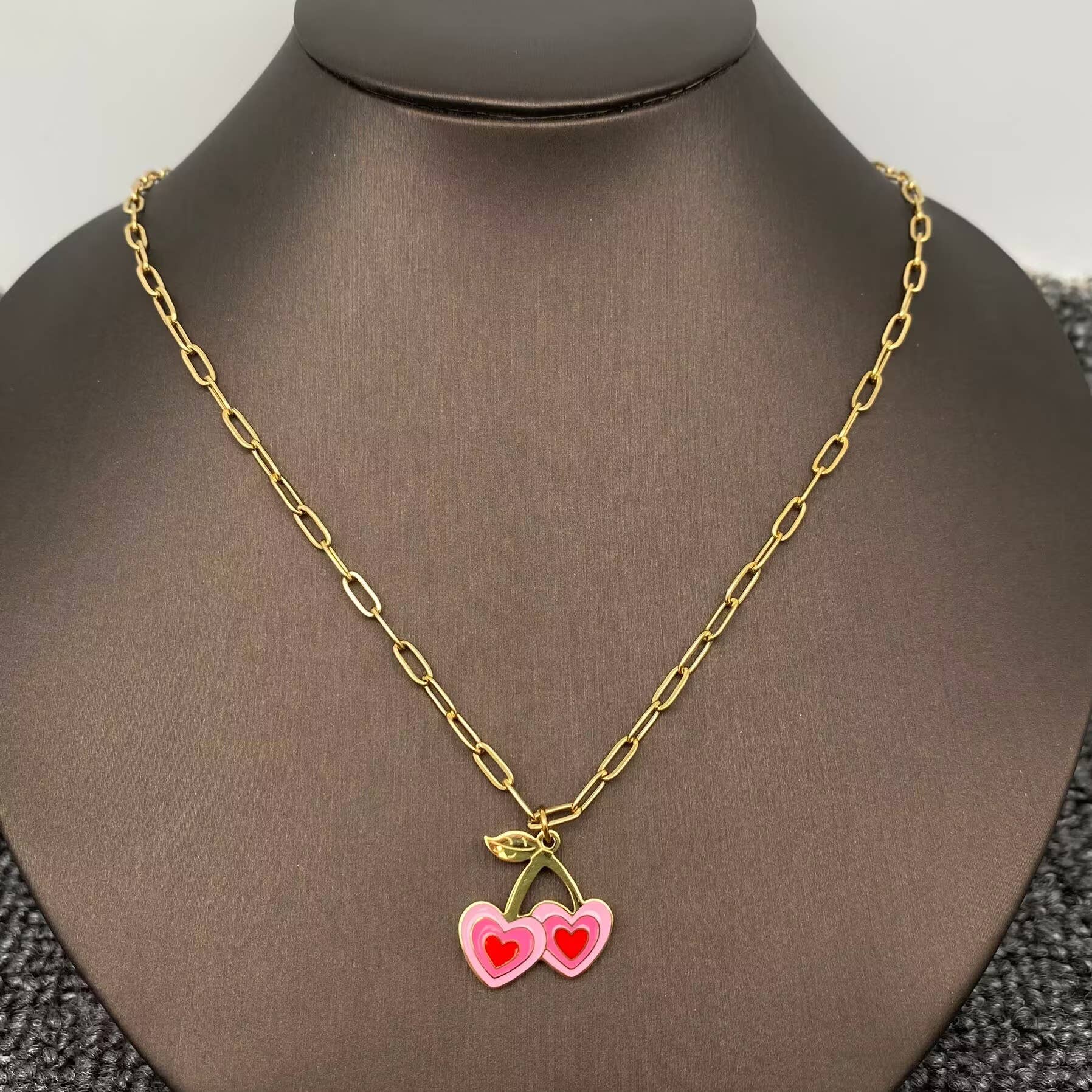 a pink and red cherry themed enamel necklace with gold chain on a black necklace stand