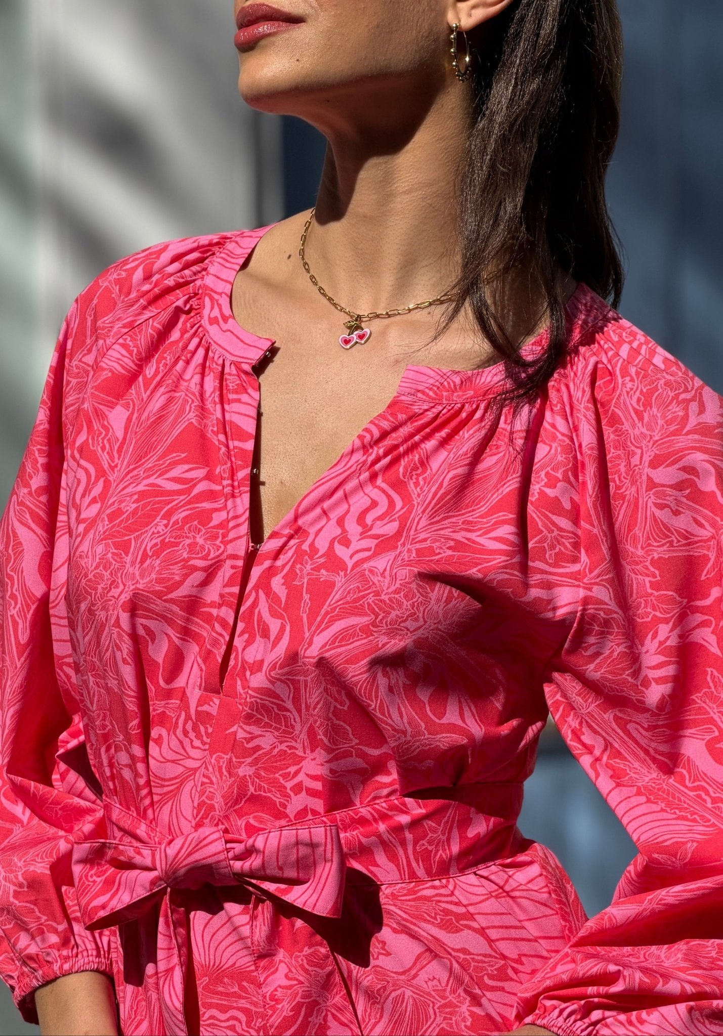 Woman wearing a pink patterned dress and a cherry heart necklace with a blurred background