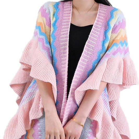 A woman wearing a multicolored knit poncho with horizontal stripes and tassel accents at the corners, paired with a black dress and accessorized with a golden necklace against a white background