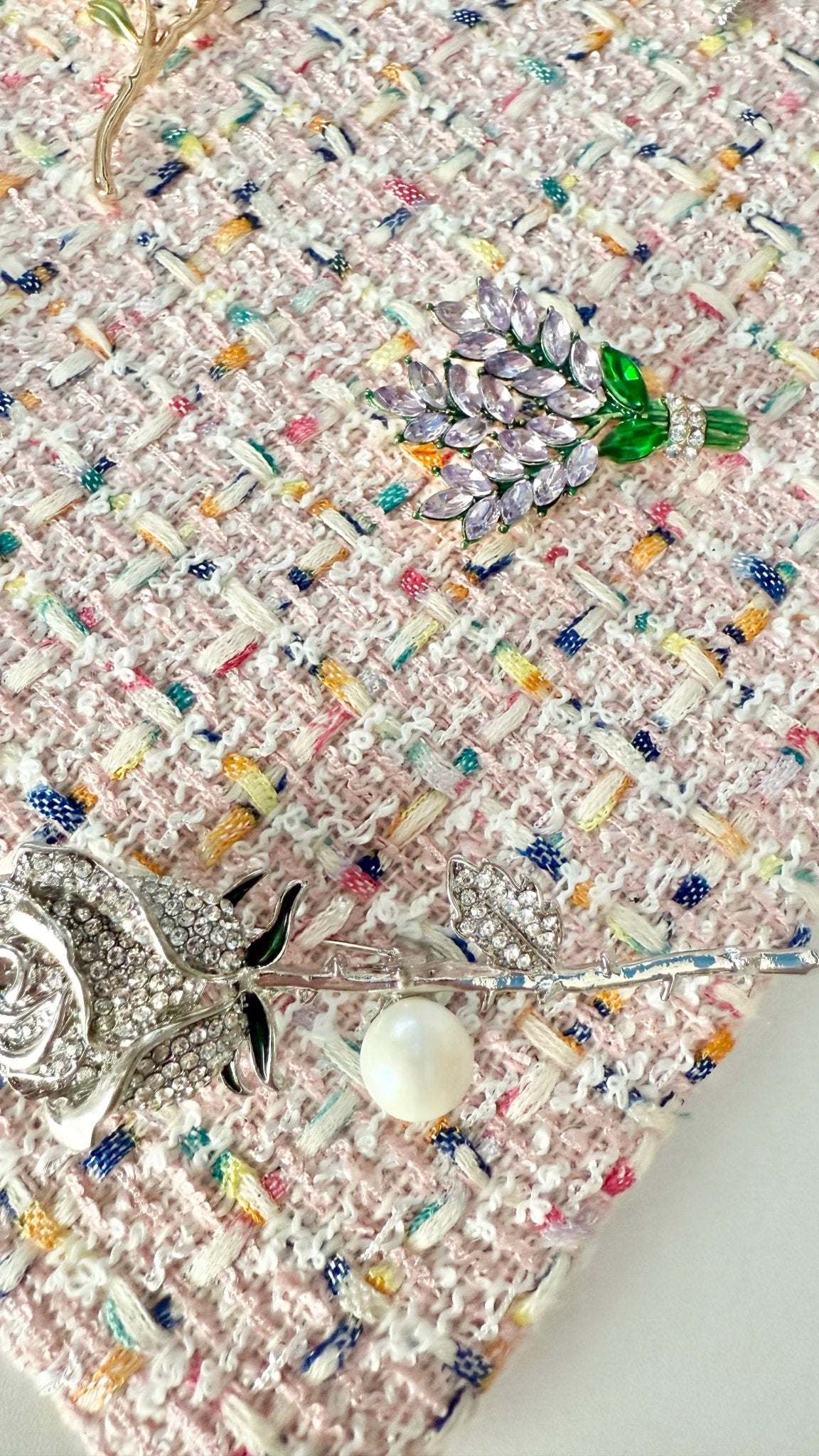 Detail view of nature-inspired jeweled brooches including lavender, rose stem with pearl, on pink tweed fabric