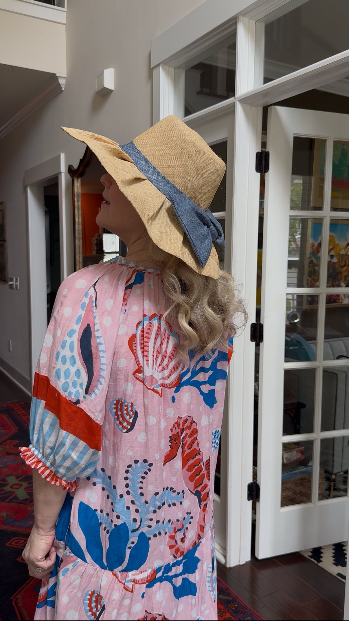 a woman with blonde hair and pink and blue ocean themed dress with tan pleated raffia sun hat with blue raffia bow