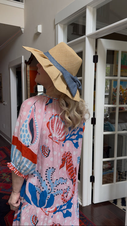 a woman with blonde hair and pink and blue ocean themed dress with tan pleated raffia sun hat with blue raffia bow