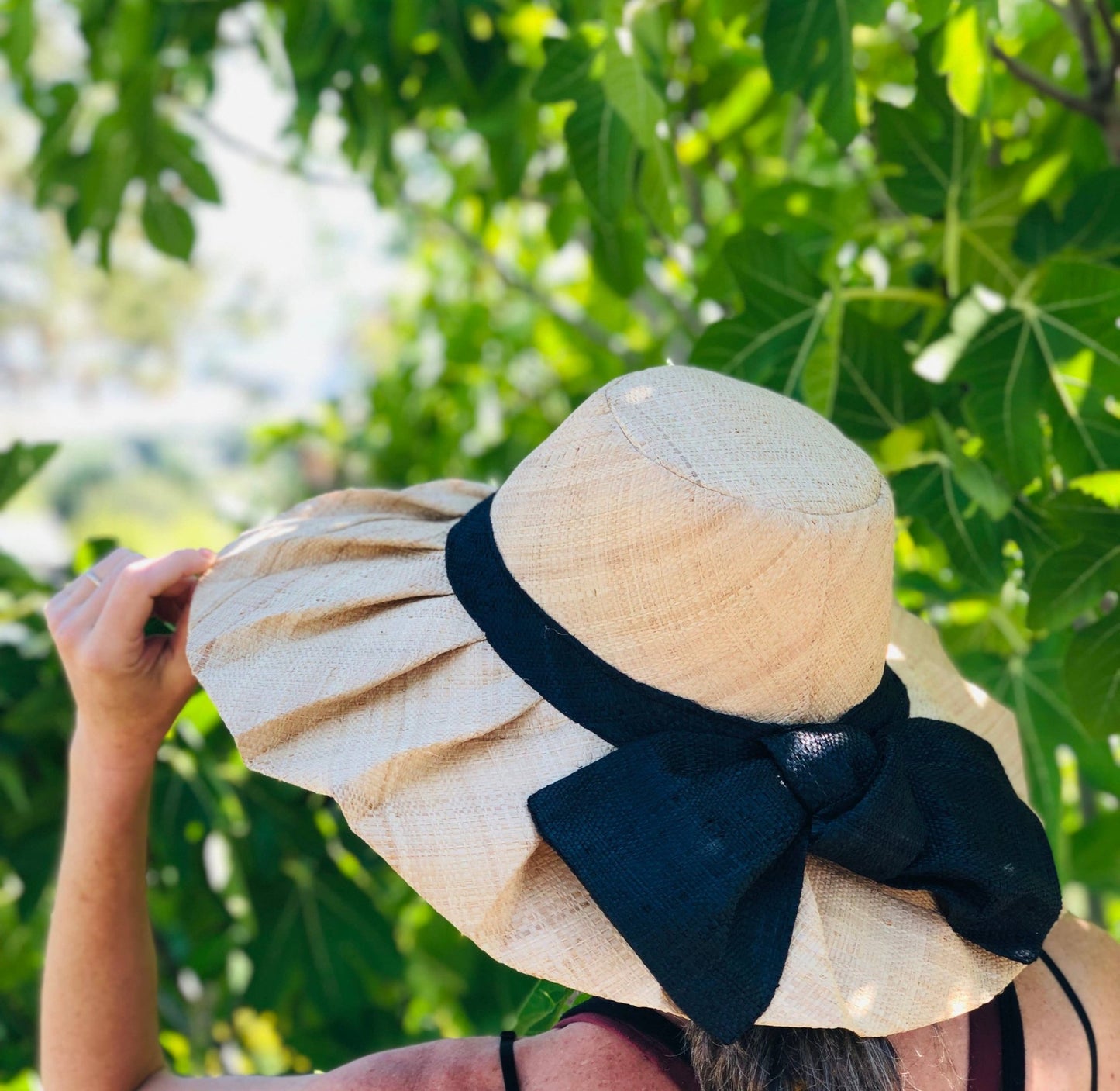 Beige pleated raffia sun hat with a black bow against a green leafy background