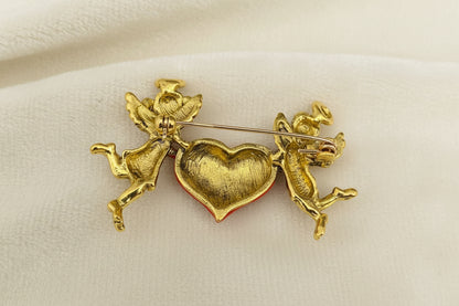 Back view of gold brooch with two cherubs holding a heart to show pin closure on a gray background