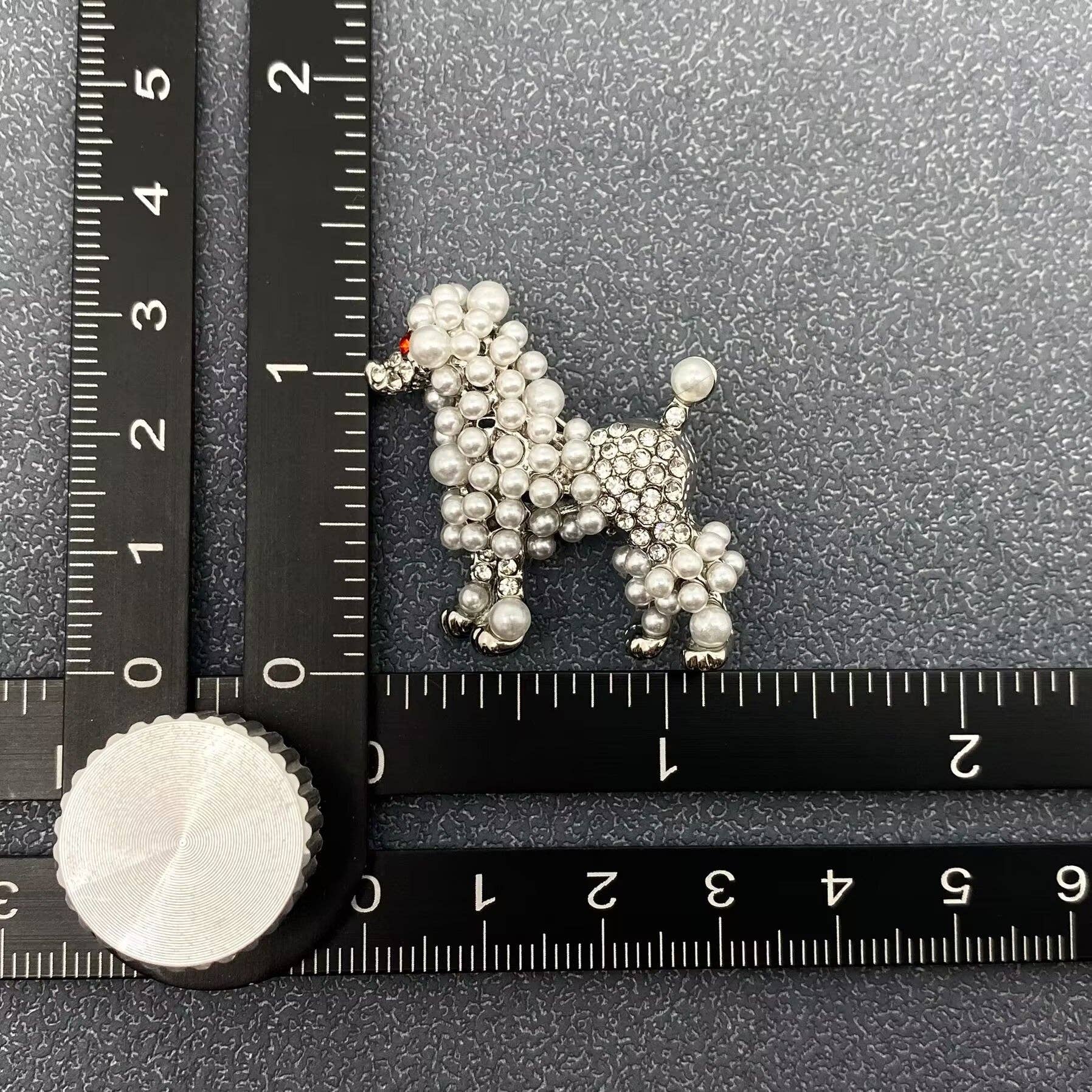 Silver poodle-shaped brooch with pearl and rhinestone embellishment on a textured black surface with a ruler for scale.