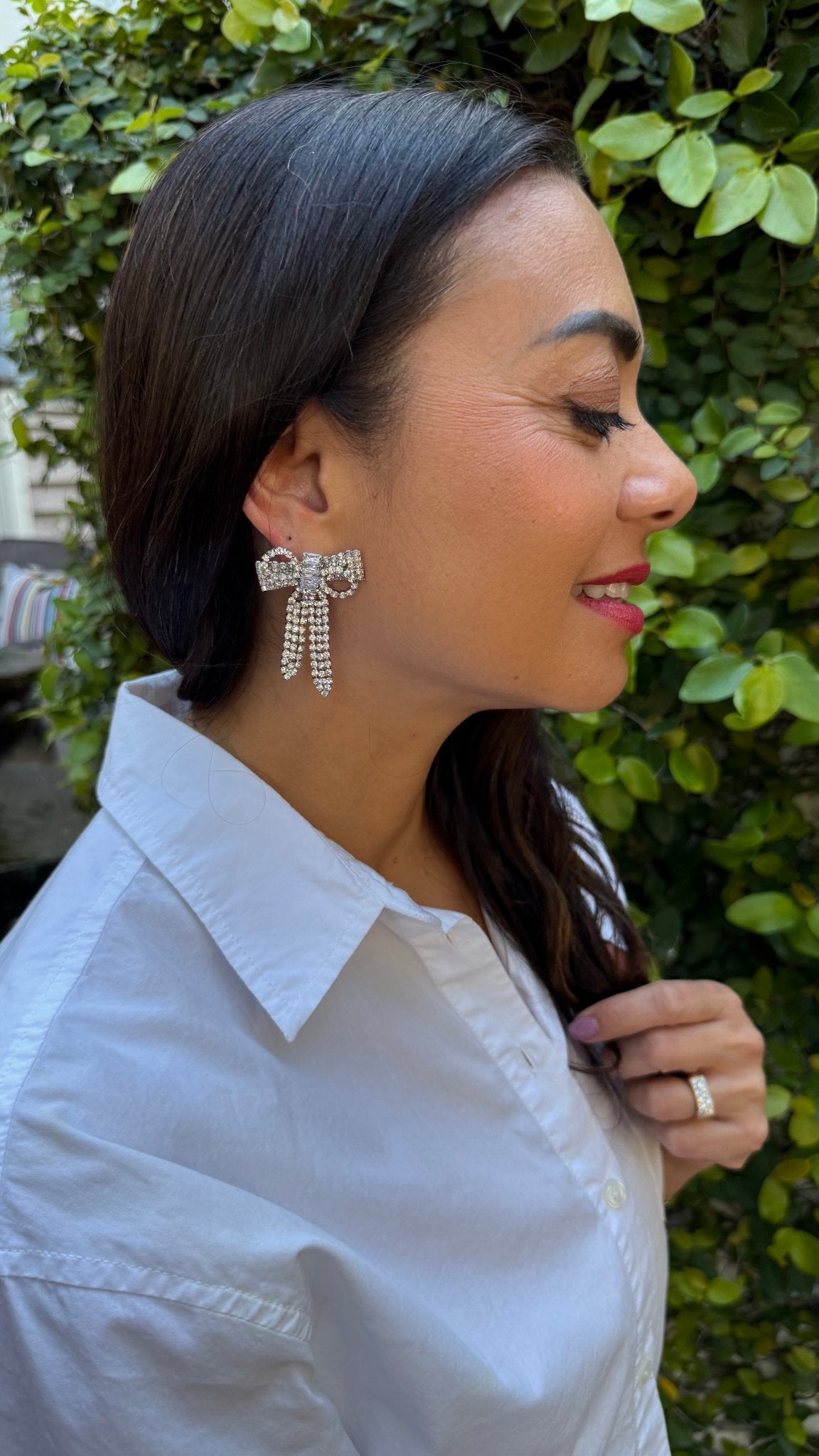 Woman wearing large bow shaped rhinestone earrings with a green leafy background