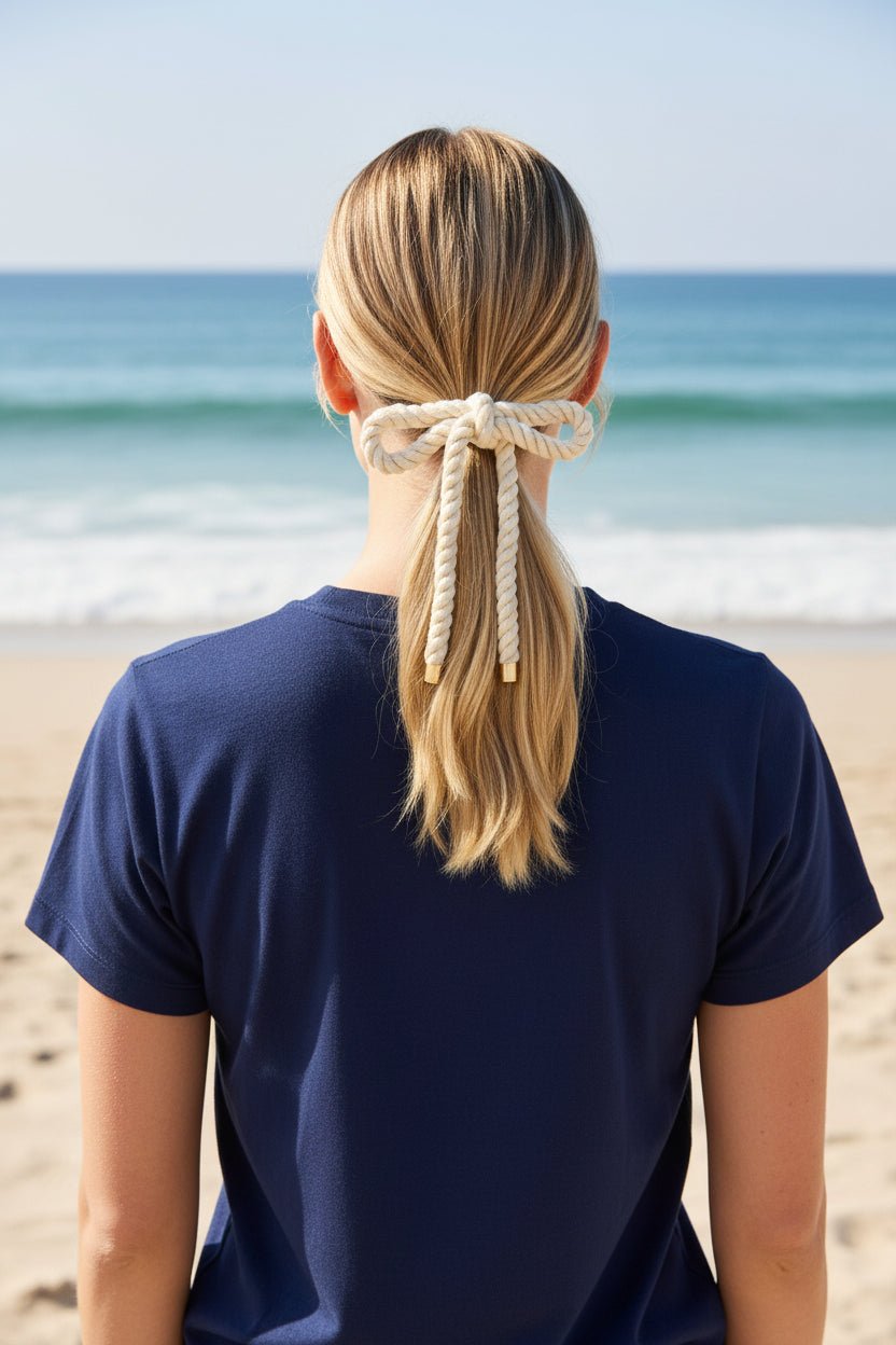 Back view of a woman wearing a beige rope bow hair clip and navy tee with the ocean in the background