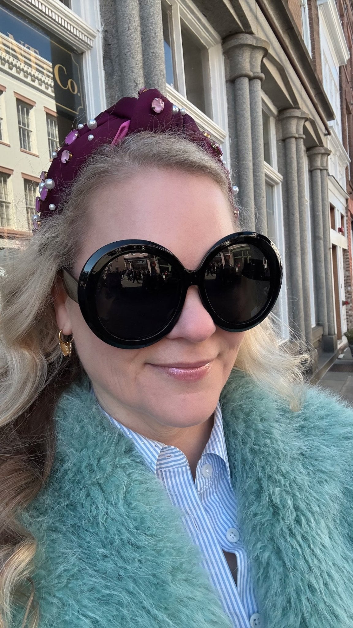 Woman wearing black round sunglasses and a teal coat in front of a building