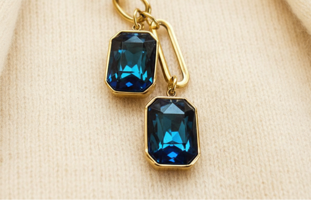 Up close view of a gold-plated stainless steel chain necklace with a double drop of faceted sapphire-colored cubic zirconia stones against an ivory sweater material background