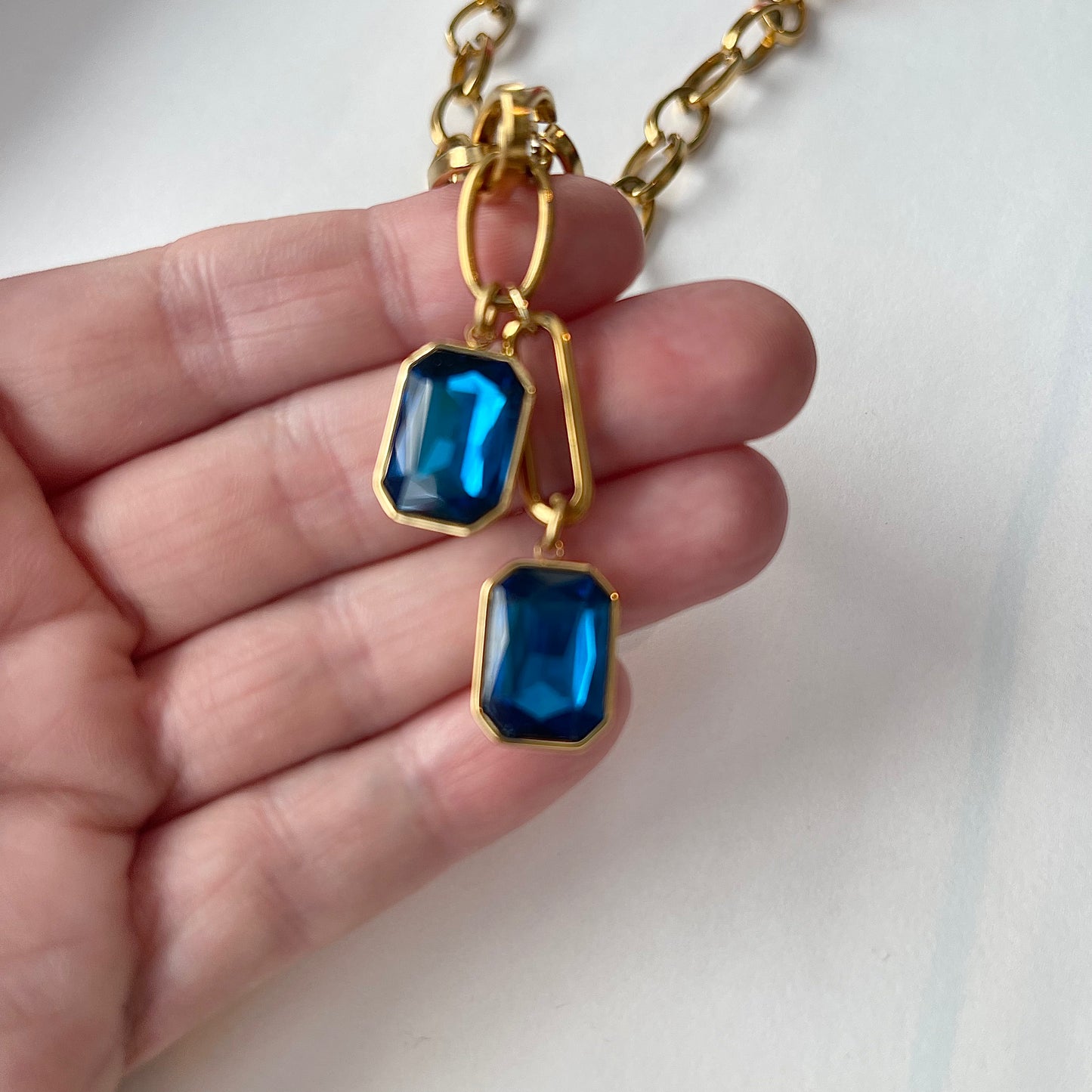 A hand holding a gold chain with two sapphire colored crystal pendants 