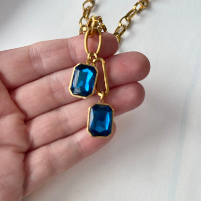 A hand holding a gold chain with two sapphire colored crystal pendants 